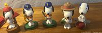 Six Snoopy character figure toys displayed on a table, each with unique costumes and poses, bases are brown, green, or white colored.