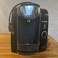 Front view of the Bosch Tassimo coffee maker showing the power cord, control button, water reservoir, and drip tray with Tassimo logo.