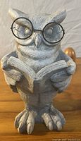 Front view of ceramic owl decor featuring the owl wearing black round glasses while holding and reading an open book.