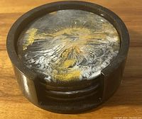 Photo of the set of four round coasters in a black circular holder featuring an abstract gold, white, and black swirl design on coaster surfaces