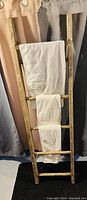 Wooden ladder towel holder with two white towels draped on rungs, photographed against curtains and a black mat.