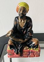 Front and side angled view of Royal Doulton Mendicant figurine showing seated man with colorful turban and black robe on red cushion