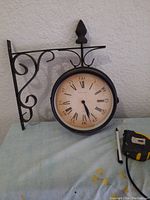 Clock showing Roman numerals on cream dial side with black metal bracket and scrollwork against textured background