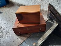 Two stacked wooden jewelry boxes; smaller one on top with light smooth finish, larger one on bottom has metal corner strips.