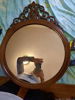 Front view of the round wood mirror showing the reflective glass and ornate carved wooden top frame detail.