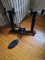 Black Soozier bicycle spin stand on wooden floor under heating radiator, adjustable mounting points, metal cylindrical inserts visible