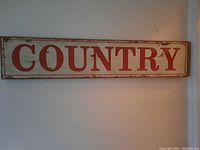 Photo of a long rectangular wooden sign with the word 'COUNTRY' painted in large red letters on a white background with distressed edges.