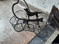 Photo showing a wire mesh basket shaped like a dog bone with a raised dog figure on top and a handle. The basket rests on a stone floor near a wall.