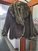 Front view of dark brown ladies faux suede jacket with large faux raccoon fur collar and faux fur trim on cuffs and hem hanging on a hanger.