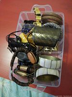 Top view of a plastic storage container with assorted bracelets including white gold accented bangles, wooden bangles, a hammered metal cuff, and other multi-strand and plastic bangles.