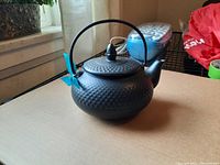 Black metal tea pot basket with hammered finish and rounded shape, curved spout, and top lid with knob. Blue ribbon tied to handle.