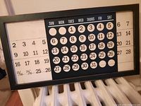 Photo of metal perpetual calendar showing days of the week and dates in black and white contrast within a rectangular frame placed on a radiator.