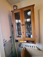 Wooden cabinet mounted on wall with two glass doors revealing glass bowls and glassware inside. Cabinet has two black handles and natural wood finish.