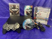 Front view of PS3 Slim console with controller, power and AV cables, Call of Duty Black Ops disc, game cases arranged next to the console covering various titles.