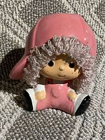 Front view of the vintage piggy bank figurine showing the child wearing a pink hat and overalls, seated with legs spread.