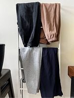 Four pairs of pants hanging on a metal rack showing colors and styles including black, dusty pink, gray, and navy blue