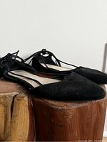 Pair of black suede flats with pointed toes and lace-up ankle straps, shown side view on a rustic wooden surface.