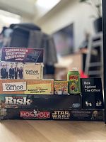 Photo showing front view of multiple board game boxes stacked and arranged. Titles visible include Codebreaker, The Office Downsizing Game, Apples to Apples Dice Game, Box Against The Office, Assistant to the Regional Manager Game, Tumbling Towers, Risk, and Star Wars Monopoly Collector's Edition.