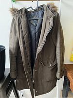 Front view of the green parka jacket, showing the faux fur trimmed hood, zipper and button closure, and flap pockets.