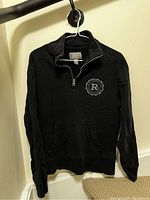 Front view of black Roots quarter zip sweatshirt hanging on hanger, showing full garment with zip and kangaroo pocket.