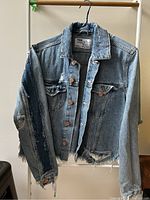 Front view of the distressed denim jacket hanging, showing frayed edges at the hem and distressed patches on sleeves, front body, and chest pockets, copper buttons visible.