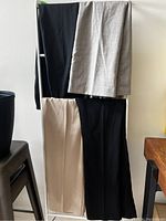 Four pairs of women's dress pants hanging on a rack: two black, one beige, and one striped gray pants.