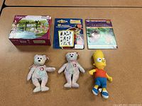 Photo showing the six items: two Ty bears, a Bart Simpson plush, boxed Schleich Bayala playset, Power Rangers tattoos booklet, and Lion King activity book.