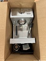 Top view of DeLonghi espresso machine inside a cardboard box showing the front, control knob, frothing wand and drip tray.