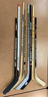 Eight assorted hockey sticks lined up on floor, showing branding and blade colors.