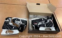 Two pairs of black and white ice hockey skates, one pair inside a box and one pair outside on table.