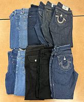 Collection of all 11 pairs folded and stacked showing fabrics and colors such as light blue, dark blue, and black denim with various pocket designs