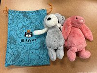 Two jellycat plush toys - a grey dog and a pink bunny - lying next to a blue Jellycat drawstring bag.