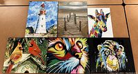 Overview of 6 paintings including lighthouse, pier, giraffe, cardinals, cat face, and bear face arranged on a table