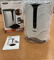 Smart pet food dispenser packaged with box and instruction manual, wrapped in plastic, showing front view with black and white design.