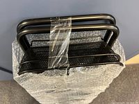 Close-up view of the top mesh shelves and tubular handles of the black metal rolling storage cart wrapped in bubble wrap.