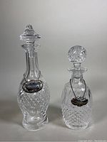 Photo showing two lidded cut crystal decanters, one taller with a round stopper and one smaller with a spherical stopper, both with silver-tone tags.