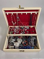 Open cream-colored jewelry box showing red velvet interior, upper section holding hanging necklaces, middle compartments with earrings and smaller jewelry pieces, bottom compartment filled with tangled necklaces and bracelets.