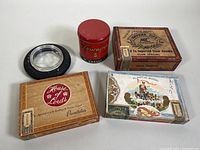 Photo showing all items together on a white surface including four empty cigar boxes, tire ashtray, and red tin.
