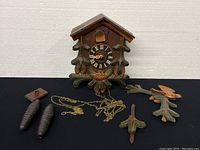 Front view of the vintage Schwarzwald cuckoo wall clock body and detached parts including pinecone weights, pendulum, and decorative bird piece.