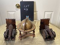 Photo showing the pair of wooden bookends shaped like cannons and the miniature globe set in front of a white cabinet door on a gold-colored fabric surface.