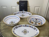 Four pieces including two decorated ceramic bowls and two matching oval serving trays with intricate floral designs and gold accents displayed on a cloth.