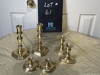 Photo showing all six brass candle holders arranged on a beige cloth surface in front of a door and a sign reading 'LOT #61'