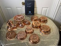 Table covered with a gold satin cloth displaying 11 assorted vintage copper molds in different shapes and sizes with embossed decorative designs, including animals and floral patterns.