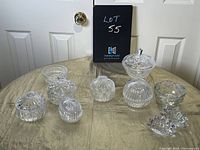 Photo of eight assorted vintage crystal and glass candy and condiment dishes arranged on a beige fabric surface with a black lot number plaque behind.