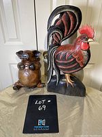 Photo showing full view of a wooden rooster painted in red, black and white standing on a base next to a light brown wooden pig with a rounded body and flat back, placed on a cloth surface in front of a door.