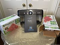 Front view of FoodSaver vacuum sealer with 3 boxes of FoodSaver sealer rolls on each side, displayed on a gold cloth with double white doors in background.