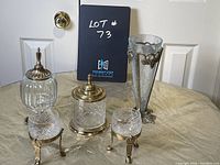 Five glass and brass decorative items arranged on a table: two covered dishes with brass lids, two condiment servers in brass stands, and a tall crackled glass vase with brass grapevine design.