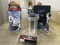 Photo of lot items including boxed Trudeau ice crusher, acrylic wine cooler, glass decanter, and boxed wine stoppers on table.