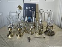 Full view of assortment of vintage brass lantern style candle holders on a beige cloth background. Includes nine items: multiple brass candle holders with glass chimneys and a brass candle snuffer.
