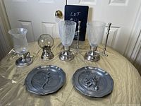 All six pewter items arranged on a table with beige cloth: two large lanterns with textured glass shades and handles, a small hurricane candle holder with glass chimney, a small globe oil lamp, and two pewter collectible plate with embossed figures.